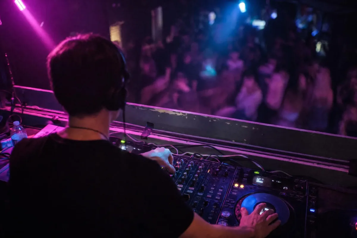 DJ working a mixer behind a booth with colored wash lighting and a blurred dance floor beyond