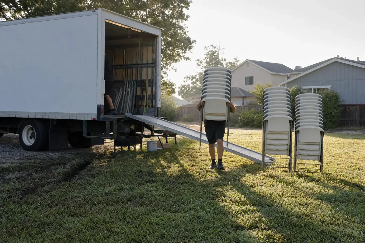 Workers unloading stacks of folding chairs and tables from a box truck onto a lawn