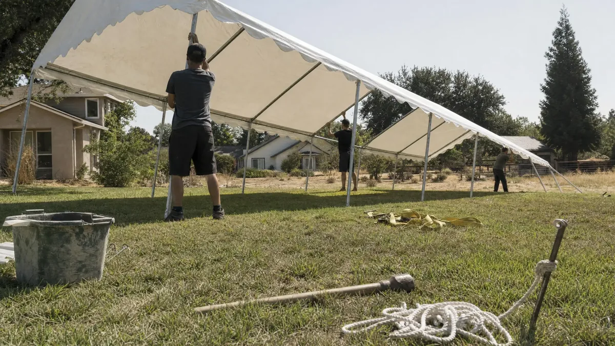 Crew raising a white frame tent on a lawn with straps and stakes on the ground